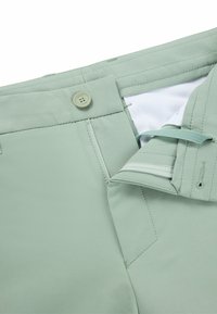 Light green trousers with a smooth texture, featuring a button closure, zip fly, and contrasting interior lining. Subtle stitching details.