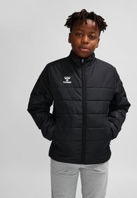 Sort polstret jakke med lynlås foran, høje krave og hvidt logo på brystet; båret over grå sweatpants. Glat tekstur, letvægtsdesign.