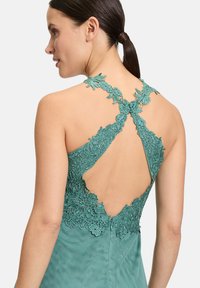 Turkusowa sukienka z koronkowym tyłem, zdobiona kwiatowymi akcentami, z v-kształtnym wycięciem i gładkim, strukturalnym wykończeniem tkaniny.