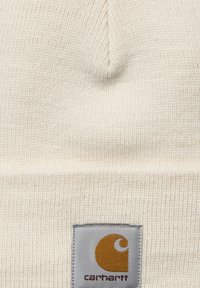 Primo piano di un berretto a maglia color crema con un'etichetta grigia cucita sopra, che presenta il logo Carhartt in marrone e arancione.