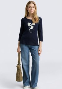 Granatowa koszula z długim rękawem w kwiatowy wzór, zestawiona z jasnoniebieskimi jeansami. Modelka trzyma beżową torebkę; casualowy krój z okrągłym dekoltem.