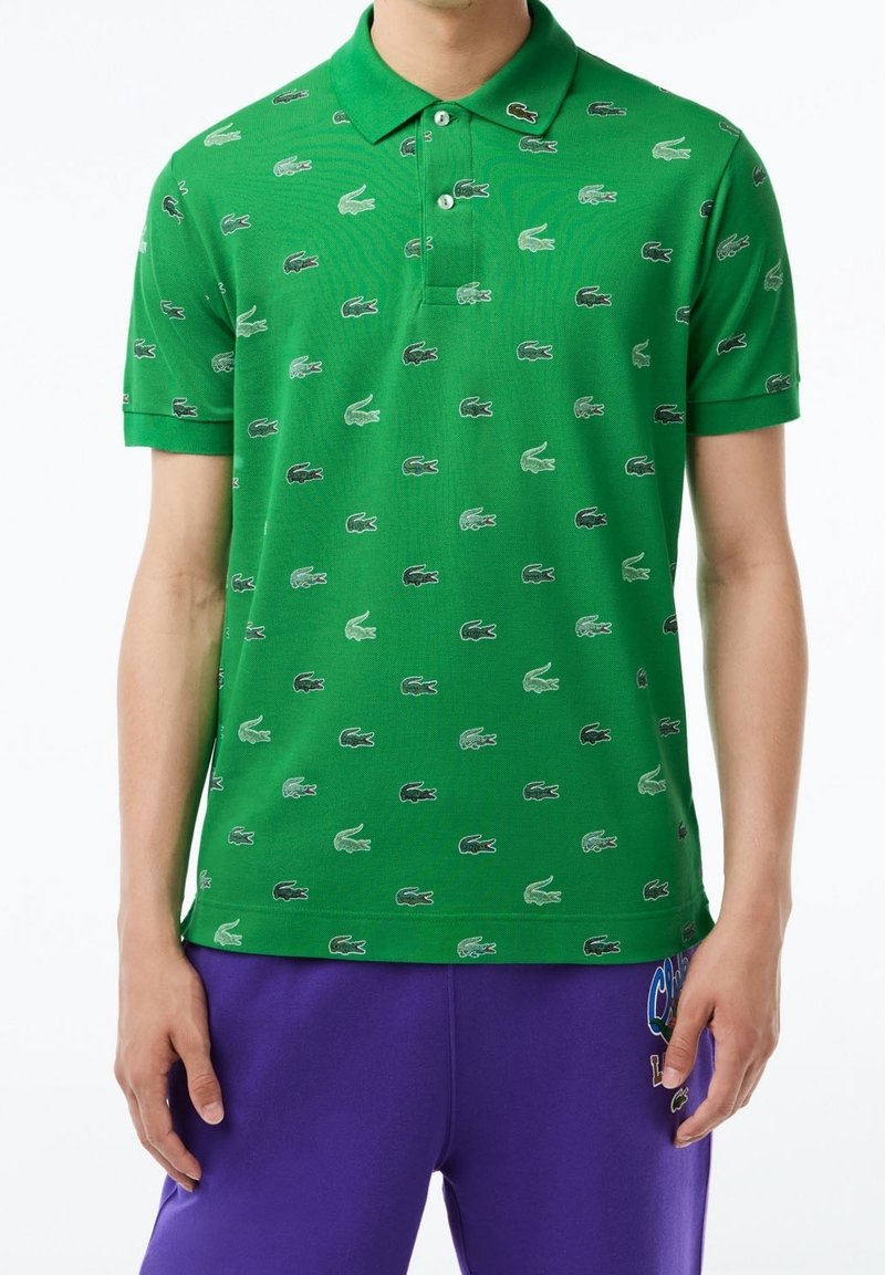 Polo vert à manches courtes avec un motif de petits logos crocodile noirs et blancs, accompagné d’un short violet avec un patch coloré.
