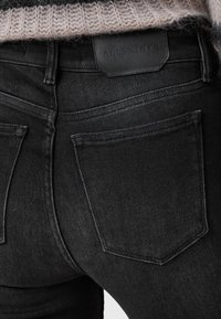 I jeans in denim nero presentano un etichetta in pelle con la scritta "ALLSAINTS" sul retro, cuciture sottili e un design aderente. Il tessuto ha una consistenza liscia.