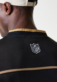 Black NFL jersey with mesh fabric, gold and black striped neckline. Features embroidered NFL logo on the back with star accents.
