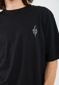 Svart bomull T-skjorte med en liten hvit slangebroderi på venstre bryst. Klassisk passform med korte ermer og rund hals.