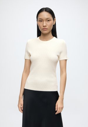 Femme aux cheveux noirs et lisses portant un haut en tricot crème à manches courtes et une jupe noire, debout devant un fond clair uni.