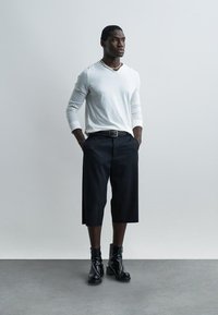 Άντρας που στέκεται με τα χέρια στις τσέπες, φοράει λευκό πουκάμισο με μακριά μανίκια, μαύρα φαρδιά σορτς μέχρι το γόνατο, μαύρες μπότες και περιδέραιο με πέρλες, σε γκρι φόντο.