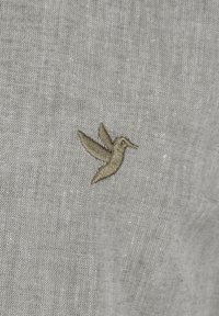 Diseño de colibrí bordado en beige sobre una tela de lino gris claro, con una superficie texturizada y ligeramente tejida.