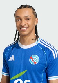 Blaues Sporttrikot mit weißen Akzenten, das einen runden Kragen und ein Logo aufweist. Das Material wirkt leicht mit einer glatten Textur und einem subtilen Muster.