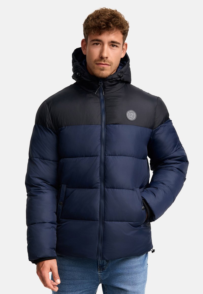 Navy blue padded jacket with a black color-blocked top section, zip closure, hood, and logo detail on the chest; features side pockets.
