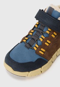 Chaussure de plein air bleue et marron avec une tige en daim et cuir, une sangle Velcro et des accents jaunes sur le système de laçage et la semelle.