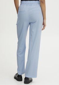 Svetlo modre hlače širokega kroja z elastičnim pasom, dvema zadnjima žepoma in gladko teksturo, v kombinaciji s črnimi čevlji.