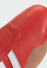 Crvene kožne cipele s glatkom teksturom, kontrastnim bijelim prugama i zaobljenim vrhom. Uključuju detalje šivanja duž rubova.