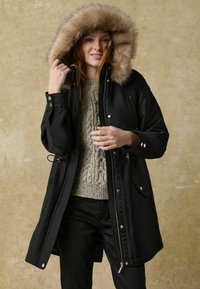 Parka noire avec une capuche en fausse fourrure, dotée d'une fermeture éclair, de deux poches avant et d'un pull en maille grise texturée en dessous.