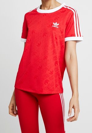 Person trägt rotes Adidas-T-Shirt und passende rote Hose mit weißen Streifen an Ärmeln und Seiten, steht vor einfachem Hintergrund.