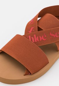 Sandale marron à enfiler avec larges brides élastiques, présentant le texte rouge "Chloé" sur les brides croisées et une semelle plate.