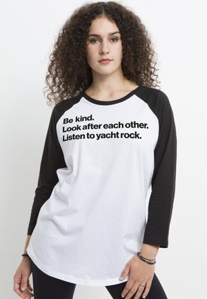 Biała koszulka z czarnymi rękawami raglanowymi z wyraźnym napisem: „Bądź miły. Troszczcie się o siebie nawzajem. Słuchaj yacht rock.” Miękki bawełniany materiał.