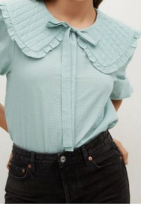 Blusa di un azzurro chiaro con texture, dotata di un ampio colletto a pieghe imbottito legato a fiocco, abbinata a jeans neri a vita alta.