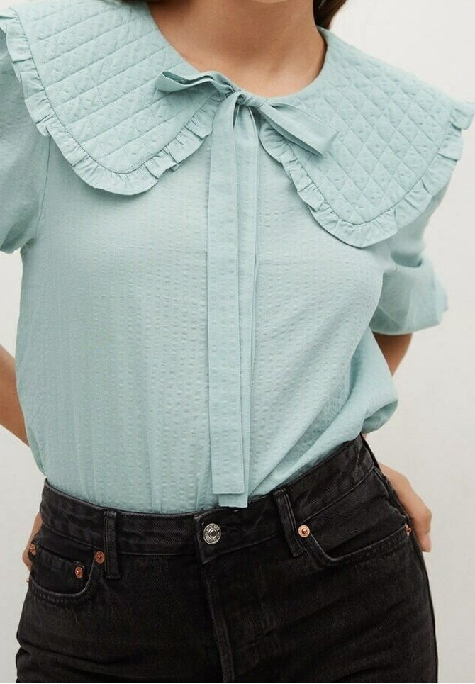 Blusa di un azzurro chiaro con texture, dotata di un ampio colletto a pieghe imbottito legato a fiocco, abbinata a jeans neri a vita alta.