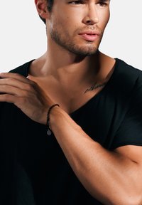 Svart flettet armbånd med rund charm, båret på håndleddet. Modellen har på seg en svart V-hals T-skjorte, som viser frem smykket og detaljene på armen.