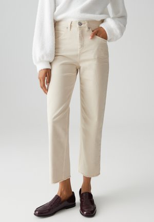 Person iført cremefarvede cropped jeans med frynset kant, hvid fluffy sweater og mørkerøde loafers, stående mod grå baggrund.