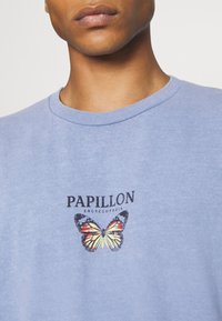 Světle modré bavlněné tričko s kulatým výstřihem, na kterém je grafika motýla a text "PAPILLON ENCYCLOPEDIA" vytištěný pod ním.