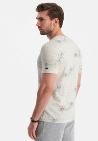 Camiseta crema con un patrón texturizado de puntos negros dispersos, mangas cortas y una etiqueta de tela negra en la manga izquierda.