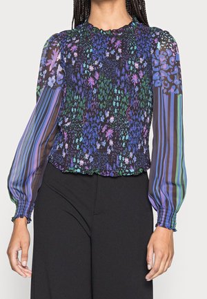 Kvinde iført en bluse med lange ærmer med flerfarvede blomster- og stribede mønstre og sorte bukser, stående med afslappede arme.