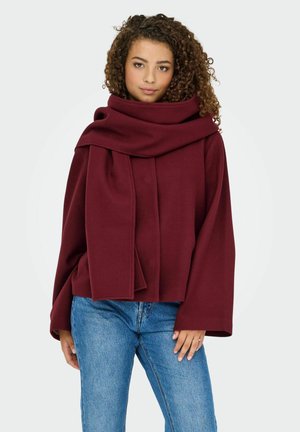 Bordeaux oversized jasje met een gedrapeerde, sjaalachtige kraag en brede mouwen, gecombineerd met strakke blauwe jeans. Zachte textuur, geen zichtbare patronen.
