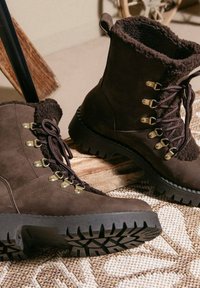 Bruine suède laarzen met faux fur voering, vetersluiting, gouden hardware oogjes, robuuste rubberen zolen en een opvallend profielpatroon.