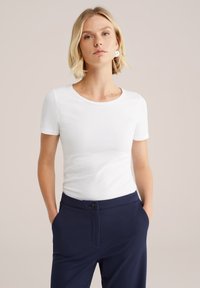 Blonde Frau mit kurzen Haaren trägt ein weißes, kurzärmliges Hemd, das in navyfarbene Hosen gesteckt ist, und steht mit den Händen in den Taschen vor einem beigen Hintergrund.