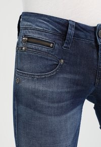 Mörkblå denimjeans med en framzip ficka, rundade sidofickor och en liten logotyp ovanför framfickan.