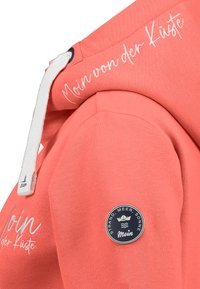 Koraal hoodie met witte trekkoord, witte scripttekst "Moin von der Küste" en een ronde patch met de tekst "Strand, Meer, Sonne Moin" op de mouw.