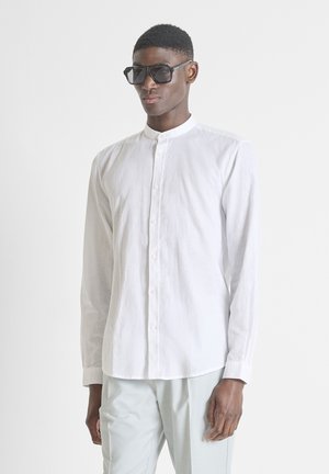 Mann mit großen schwarzen Sonnenbrillen, weißem Hemd mit Mandarinkragen und hellen Hosen, der vor einem schlichten Hintergrund steht.