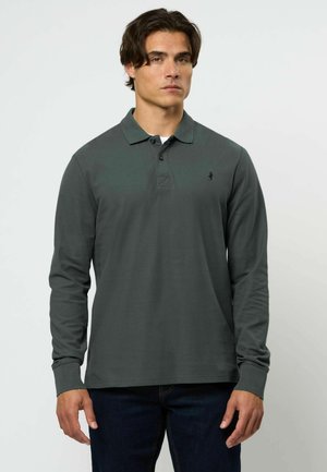 Muž v tmavozelenej dlhoruká polo košeli a tmavých džínsach stojí oproti svetlému jednoliatemu pozadiu, čelom vpred.