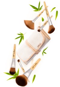 Make-up-Pinselset mit Bambusgriffen und silbernen Ferrulen, angeordnet um eine strukturierte weiße Rolltasche, begleitet von grünen Bambusblättern.