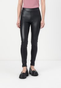 Kvinna iklädd svarta imitation läderleggings och kraftiga svarta plattformsskor, stående mot en vit bakgrund.