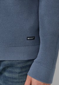 Jasnoniebieski sweter o fakturze z prążkowanymi mankietami i dołem. Posiada małą metkę "Elevated Casual" po lewej stronie, blisko dołu.