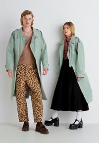 Casaco trench verde claro e calças com padrão de leopardo à esquerda; camisa xadrez e saia preta com detalhe texturizado à direita. Ambas estão a usar sapatos de plataforma.