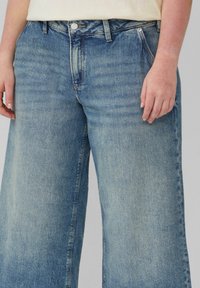 Hellblaue weite Jeans mit einem ausgewaschenen Textur, ausgestattet mit einem Knopf- und Reißverschluss sowie frontalen Taschen. Der Stoff zeigt subtile Abriebspuren.