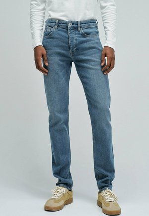 Hombre con vaqueros azul claro, zapatillas beige y camisa blanca de manga larga, de pie frente a un fondo claro y liso.