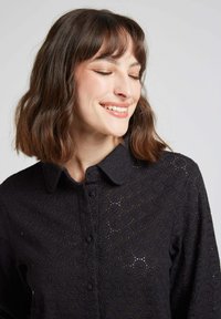 Chemisier en dentelle noire avec un design à boutons, un col et des motifs floraux circulaires sur tout le tissu. Texture douce avec des coutures visibles.