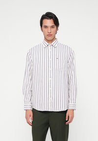 Νεαρός άντρας που φοράει ένα λευκό πουκάμισο με κάθετες μπλε και κόκκινες ρίγες και σκούρα πράσινα παντελόνια, στέκεται μπροστά σε ένα απλό λευκό φόντο.