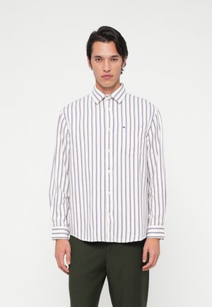 Jonge man in een wit overhemd met verticale blauwe en rode strepen en een donker groene broek, staande tegen een plain witte achtergrond.