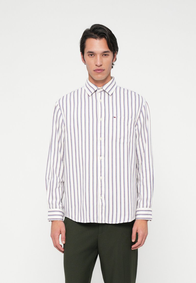 Νεαρός άντρας που φοράει ένα λευκό πουκάμισο με κάθετες μπλε και κόκκινες ρίγες και σκούρα πράσινα παντελόνια, στέκεται μπροστά σε ένα απλό λευκό φόντο.