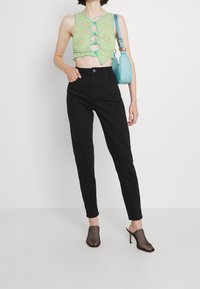 Gehaakte groene crop top met knoopaccenten, zwarte high-waisted skinny jeans, zwarte mesh open teen hakken en een lichtblauwe handtas.