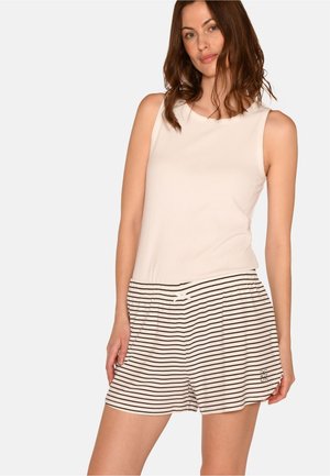 Femme portant un haut beige sans manches et un short beige avec de fines rayures horizontales noires et un petit logo près de l'ourlet.
