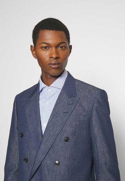 Kétgombos, sötétkék len blézer fekete gombokkal, világoskék és fehér csíkos inggel párosítva. Sima textúra.