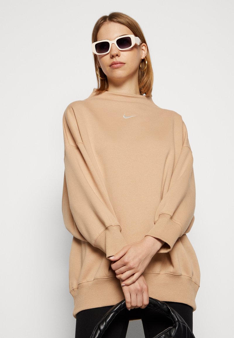 Felpa oversize beige con collo rotondo e spalle scese, con un piccolo logo Nike bianco. Il modello tiene una borsa nera.