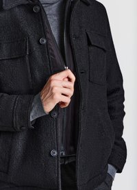 Giacca nera testurizzata con chiusure a bottone, zip frontale e due tasche sul petto. Fodera interna grigia e polsini visibili. Design semplice.